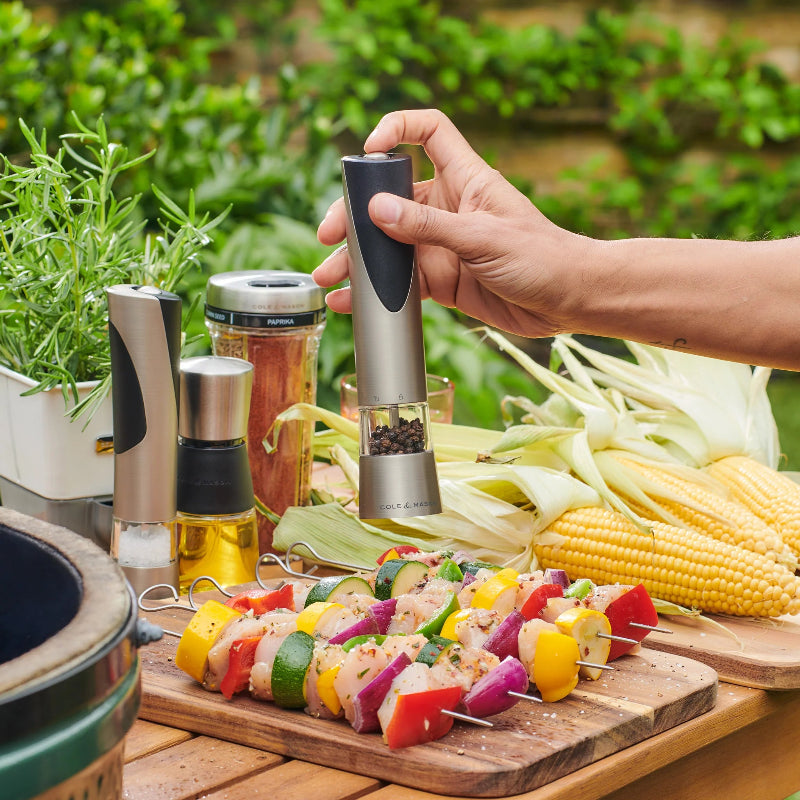 Person seasoning skewered vegetables with a pepper grinder on a wooden cutting board outdoors.