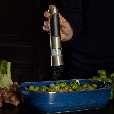 Hand using a pepper grinder over a blue dish of brussels sprouts on a dark background