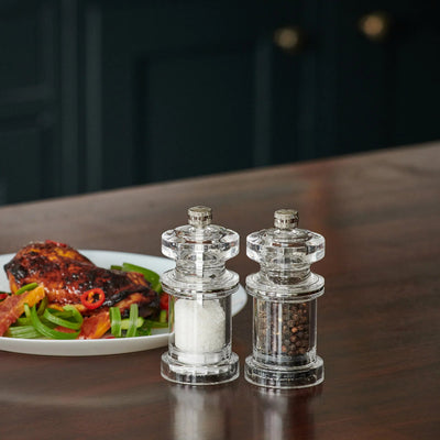 Clear salt and pepper shakers on a wooden table with a plate of food in the background.