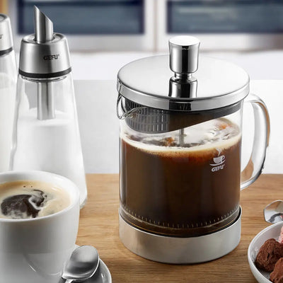 French press filled with coffee on a wooden table with a cup of coffee and pastries.