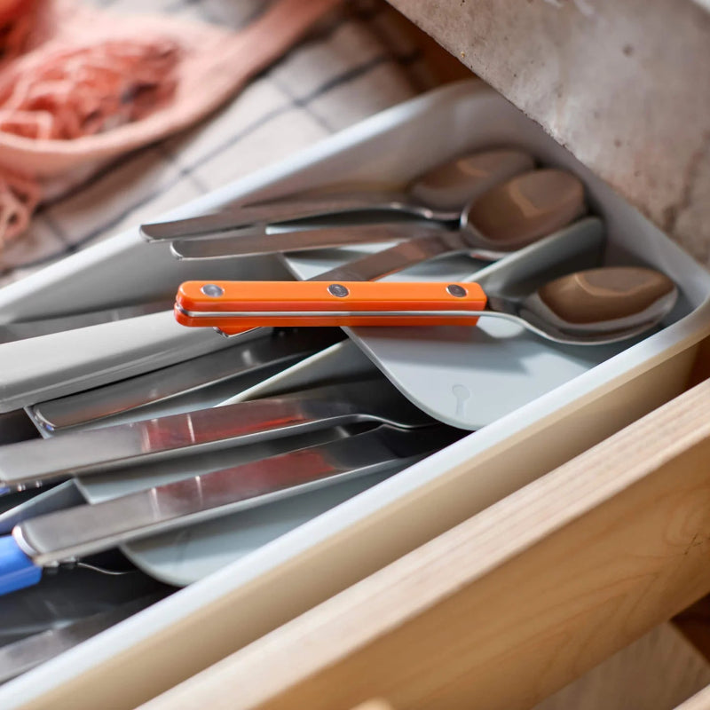 Open kitchen drawer with cutlery and an orange tool on a wooden surface.