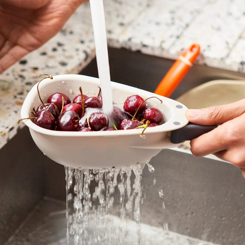 Person washing cherries in a white colander under running water in a kitchen sink.