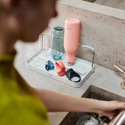 Person using a dish drying rack with various bottles on a kitchen counter.