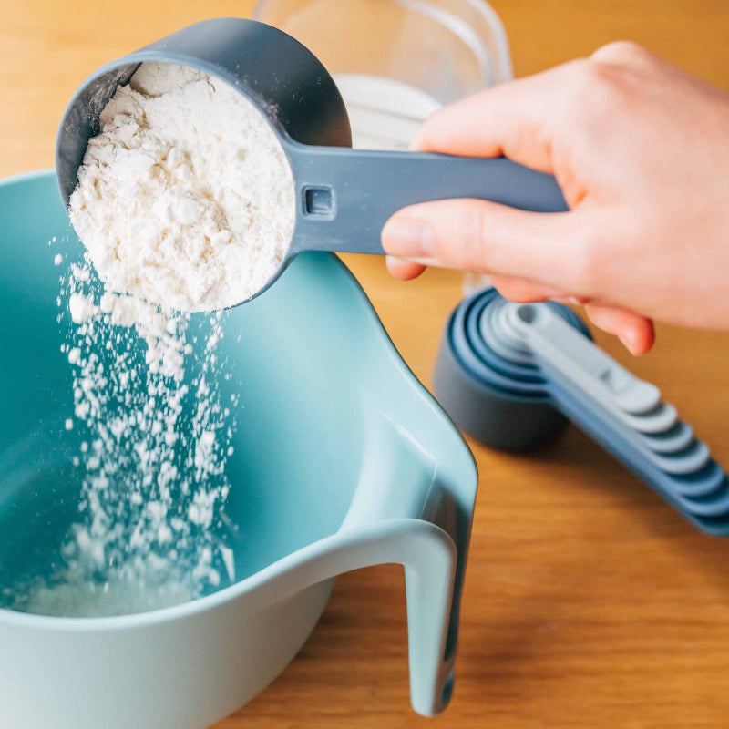 Person measuring flour into a blue bowl using a measuring cup on a wooden surface.