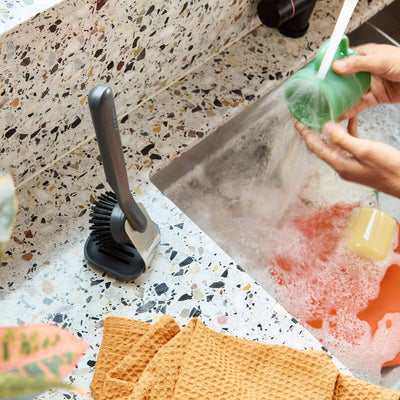 Person washing a green sponge over a sink with soapy water, on a speckled countertop.