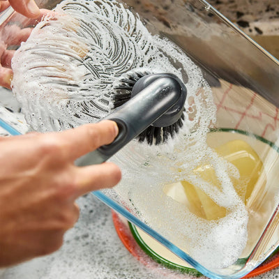 Person scrubbing a container with a brush and soapy water on a speckled countertop.