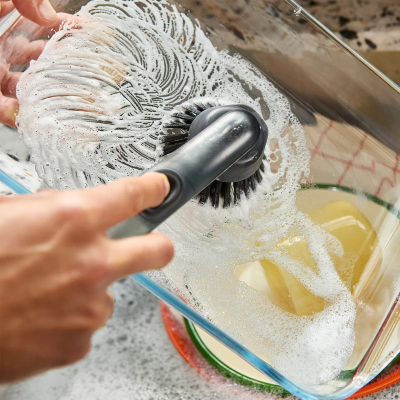 Person scrubbing a container with a brush and soapy water on a speckled countertop.