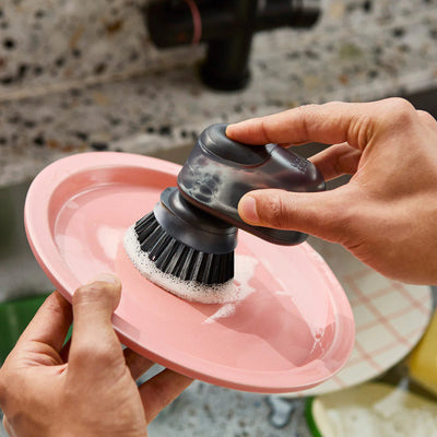 Person using a dish scrubber on a pink plate in a kitchen sink.