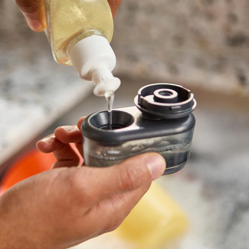 Person pouring liquid from a bottle into a small black container on a kitchen counter.