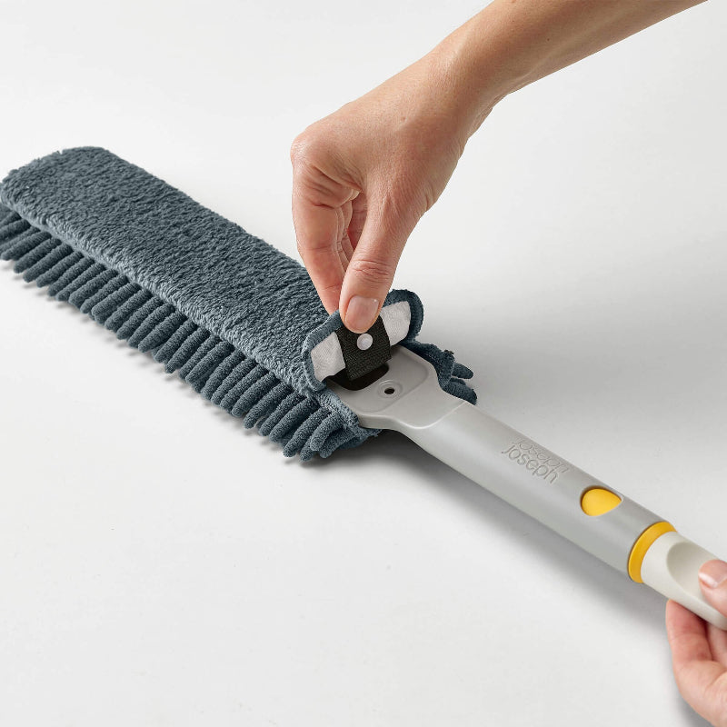 Hand holding a cleaning brush with a gray bristle head and white handle on a light background