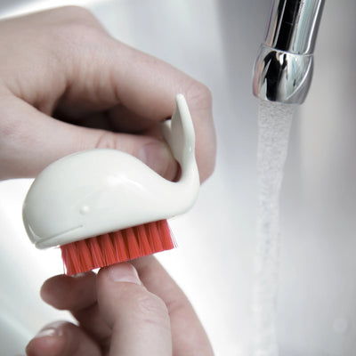 Hand holding a small white brush with red bristles under running water.