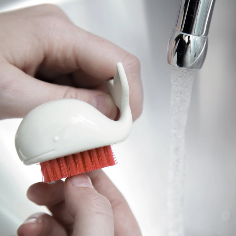 Hand holding a small white brush with red bristles under running water.