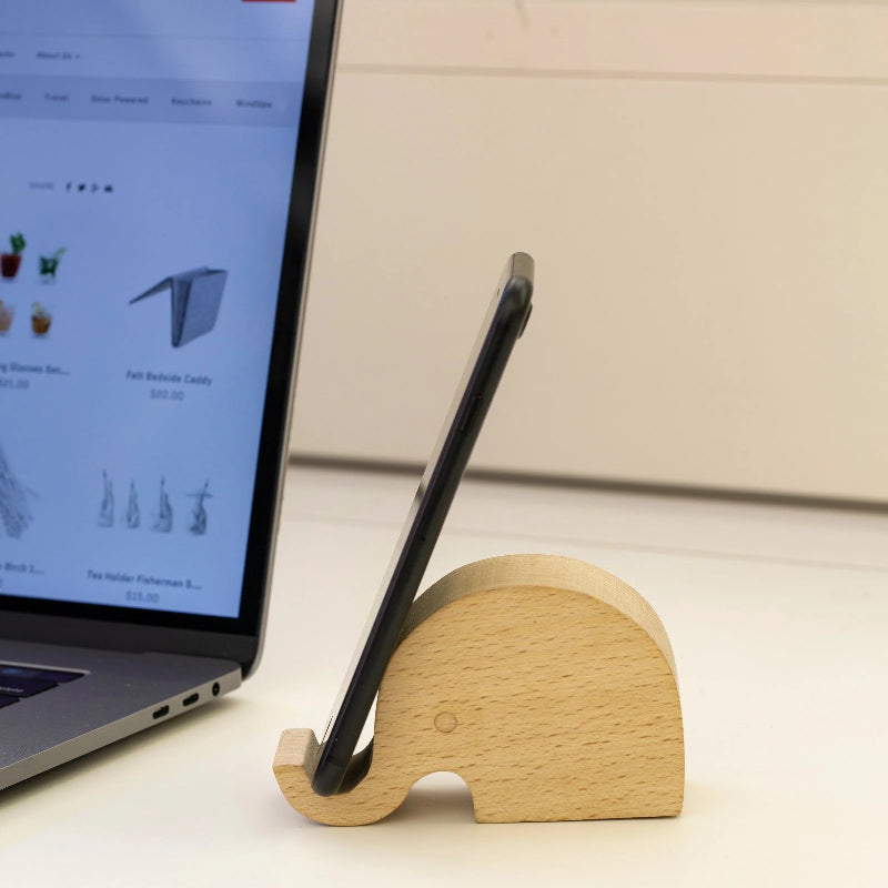 Wooden elephant phone stand holding a smartphone next to a laptop on a desk.