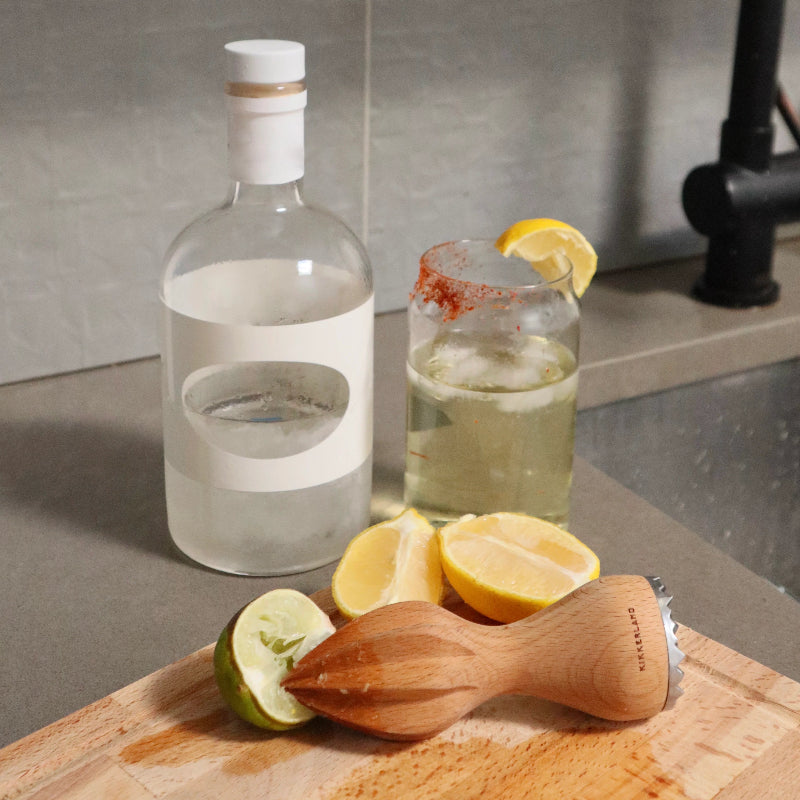 Bottle of clear liquid, glass with lemon wedge, and wooden muddler on a cutting board.