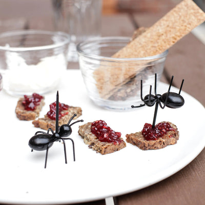 Black ant-shaped picks on crackers with jam on a white plate, with a blurred background.
