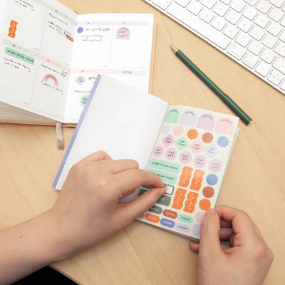 Person using colorful stickers on a planner with a keyboard and pen in the background