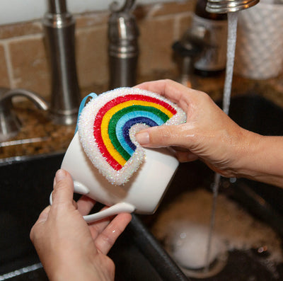 Person cleaning a mug with a rainbow sponge in a kitchen sink.