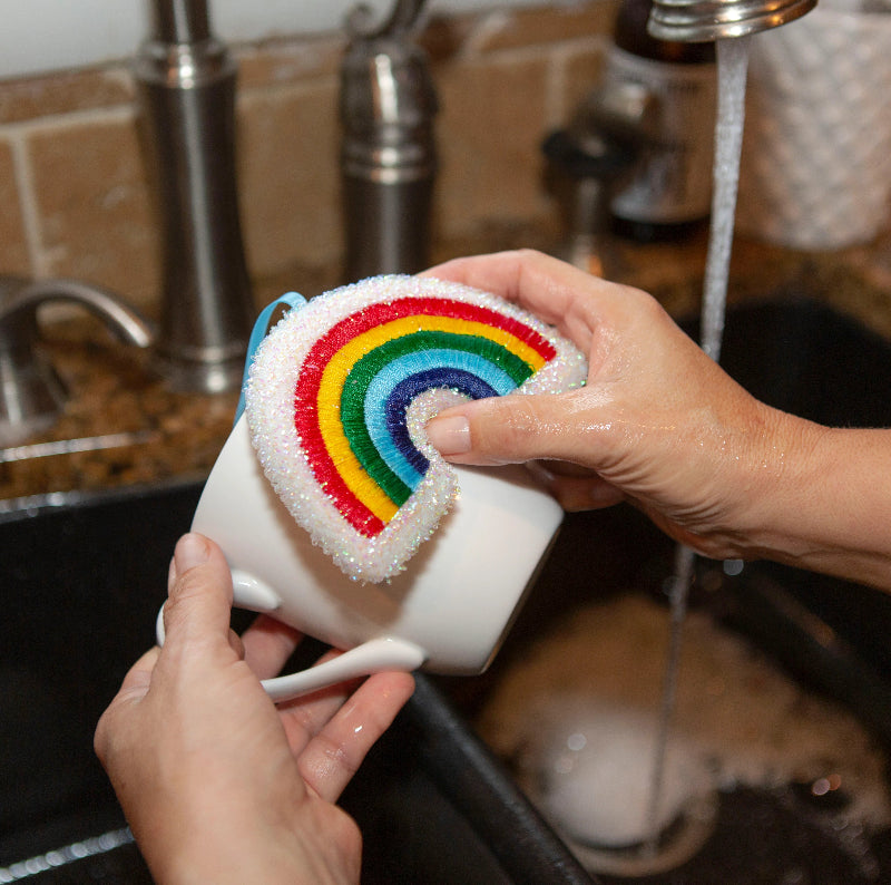 Person cleaning a mug with a rainbow sponge in a kitchen sink.
