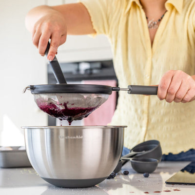 Person using a strainer to pour a berry mixture into a metal bowl on a kitchen counter.
