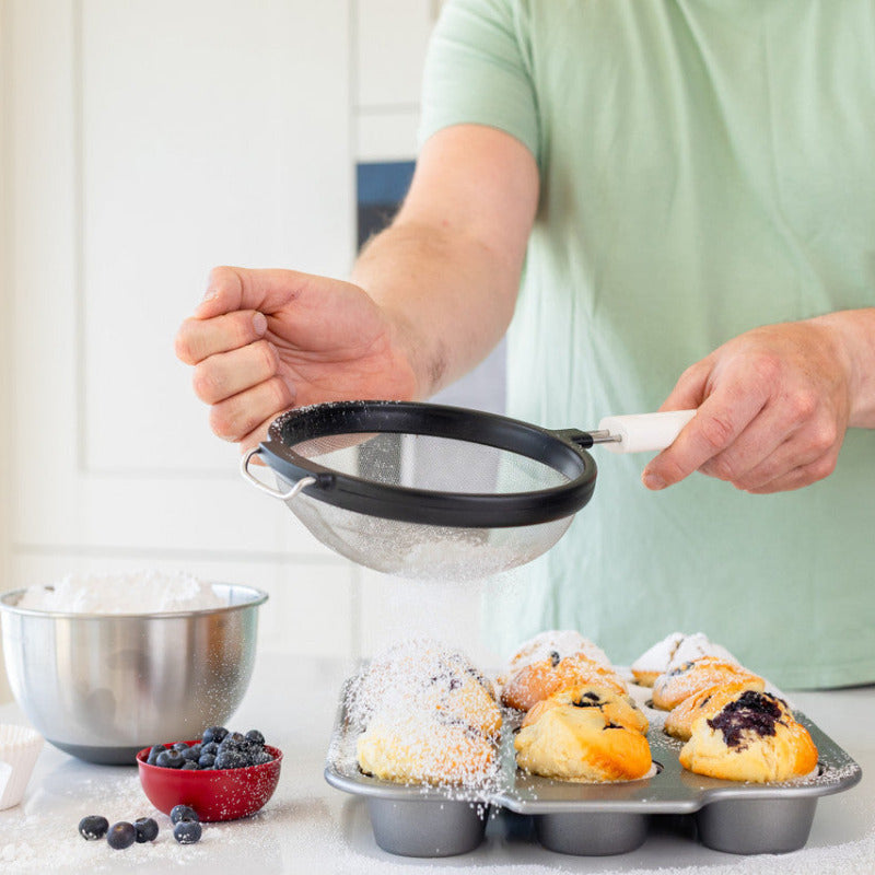 Person sifting flour into a muffin tin with baked goods on a kitchen counter.