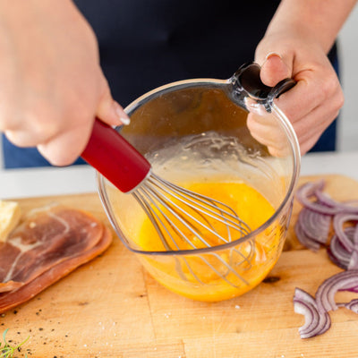Person whisking eggs in a glass bowl on a wooden cutting board with ingredients around.
