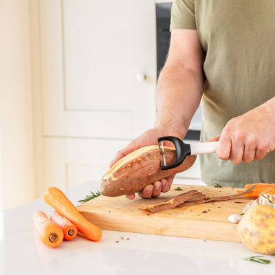 Person peeling a sweet potato on a wooden cutting board with various vegetables in a kitchen.
