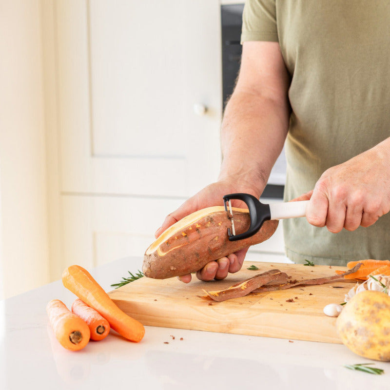 Person peeling a sweet potato on a wooden cutting board with various vegetables in a kitchen.