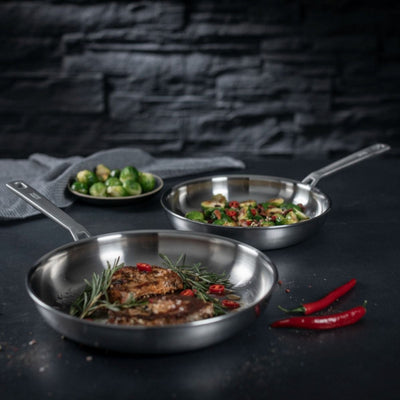 Three stainless steel frying pans with food on a dark surface and stone wall background