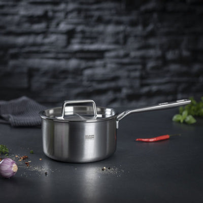 Stainless steel saucepan on a dark surface with vegetables and bread in the background