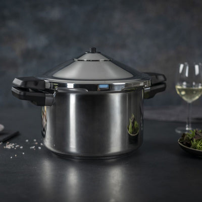 Stainless steel pressure cooker on a dark surface with wine glasses and food in the background