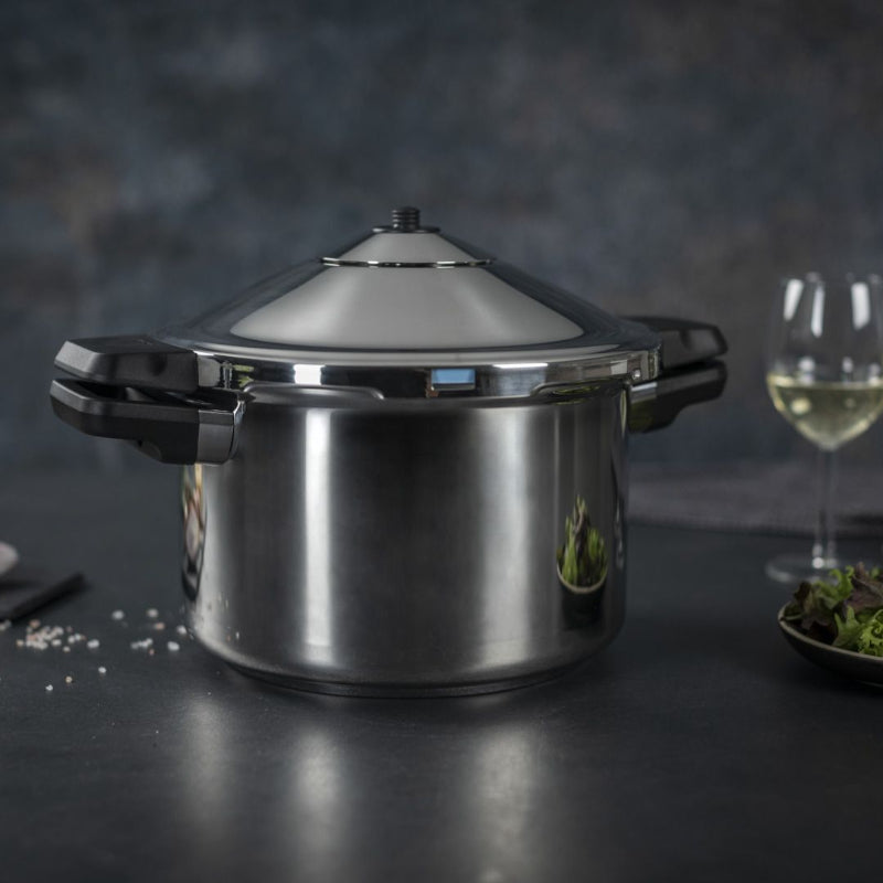 Stainless steel pressure cooker on a dark surface with wine glasses and food in the background