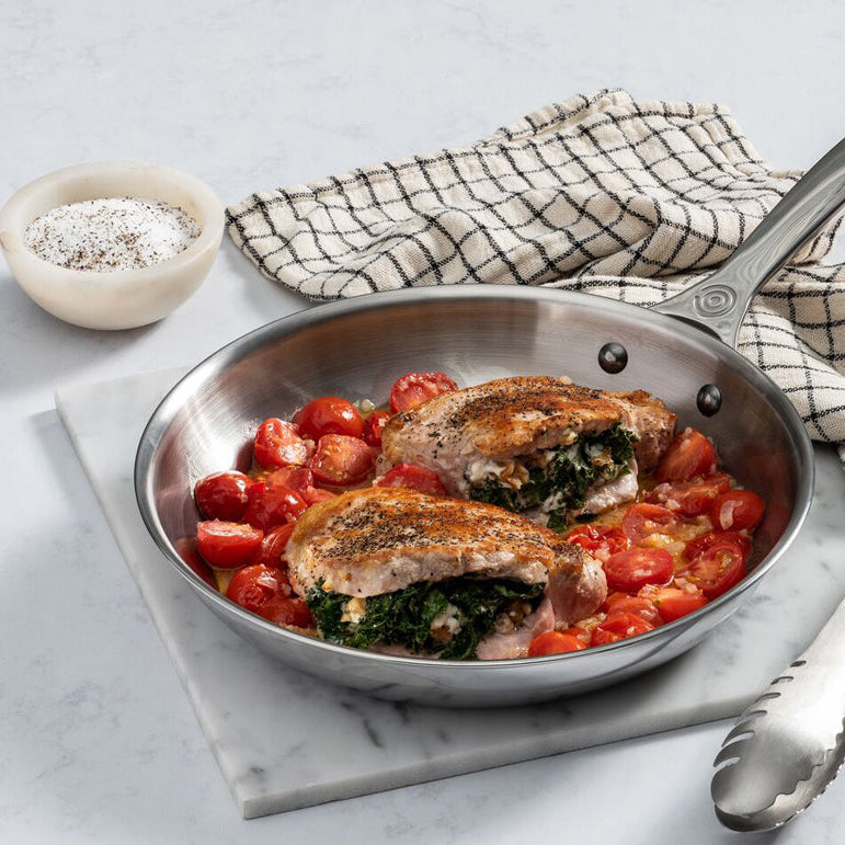 Skillet with stuffed chicken and tomatoes on a marble surface with a checkered towel.