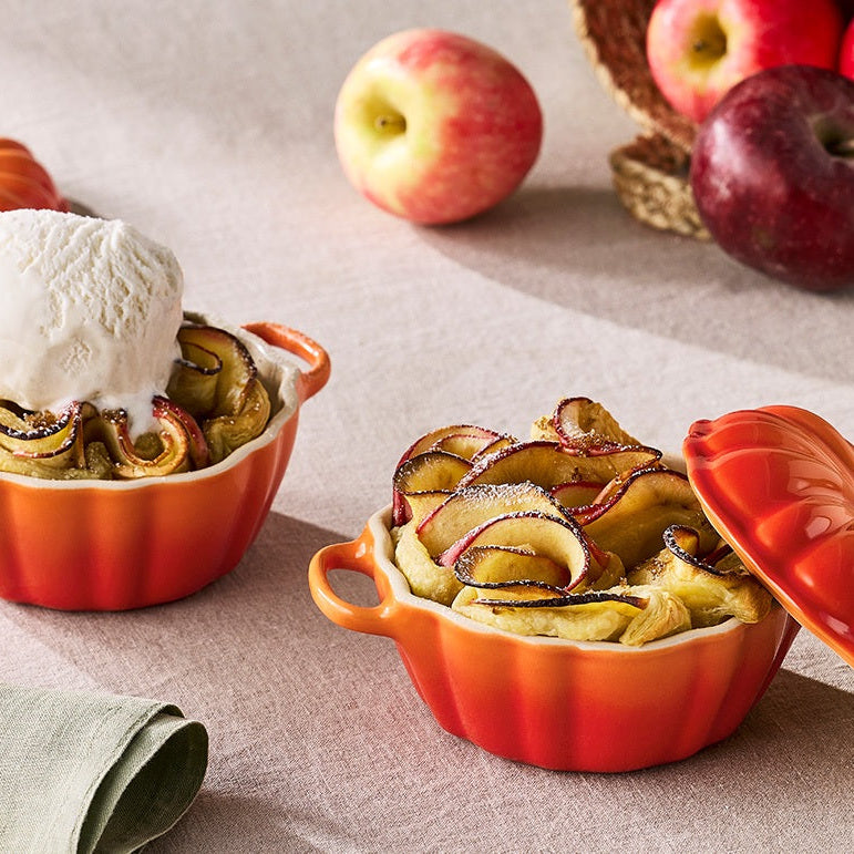 Two orange ceramic dessert dishes with apple pie and ice cream, surrounded by apples on a light background.