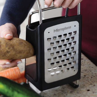 Person using a box grater to shred a potato with vegetables in the background